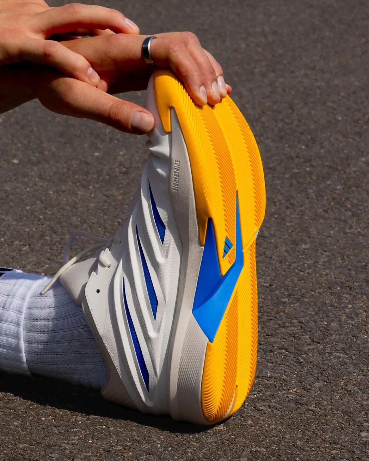 In-hand stretch showing the yellow outsole with blue insert and white upper on asphalt, Wagner Brothers