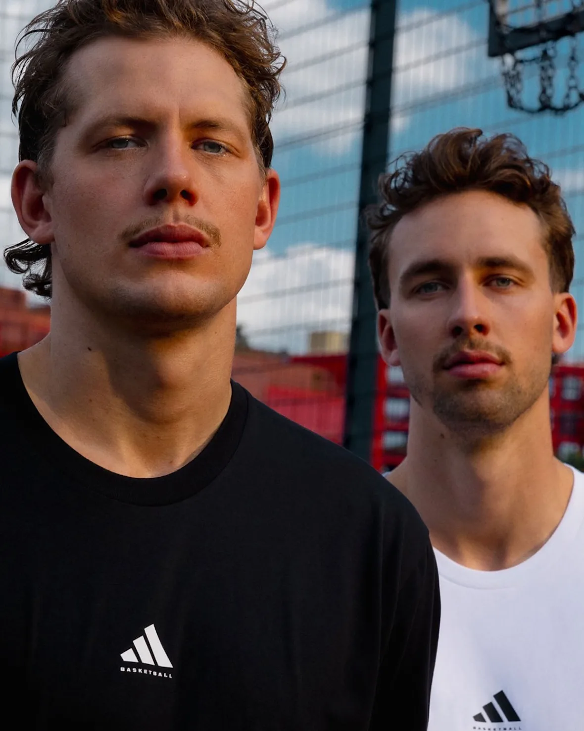 Close-up portrait of two men in black and white adidas basketball tees by a fence, Wagner Brothers