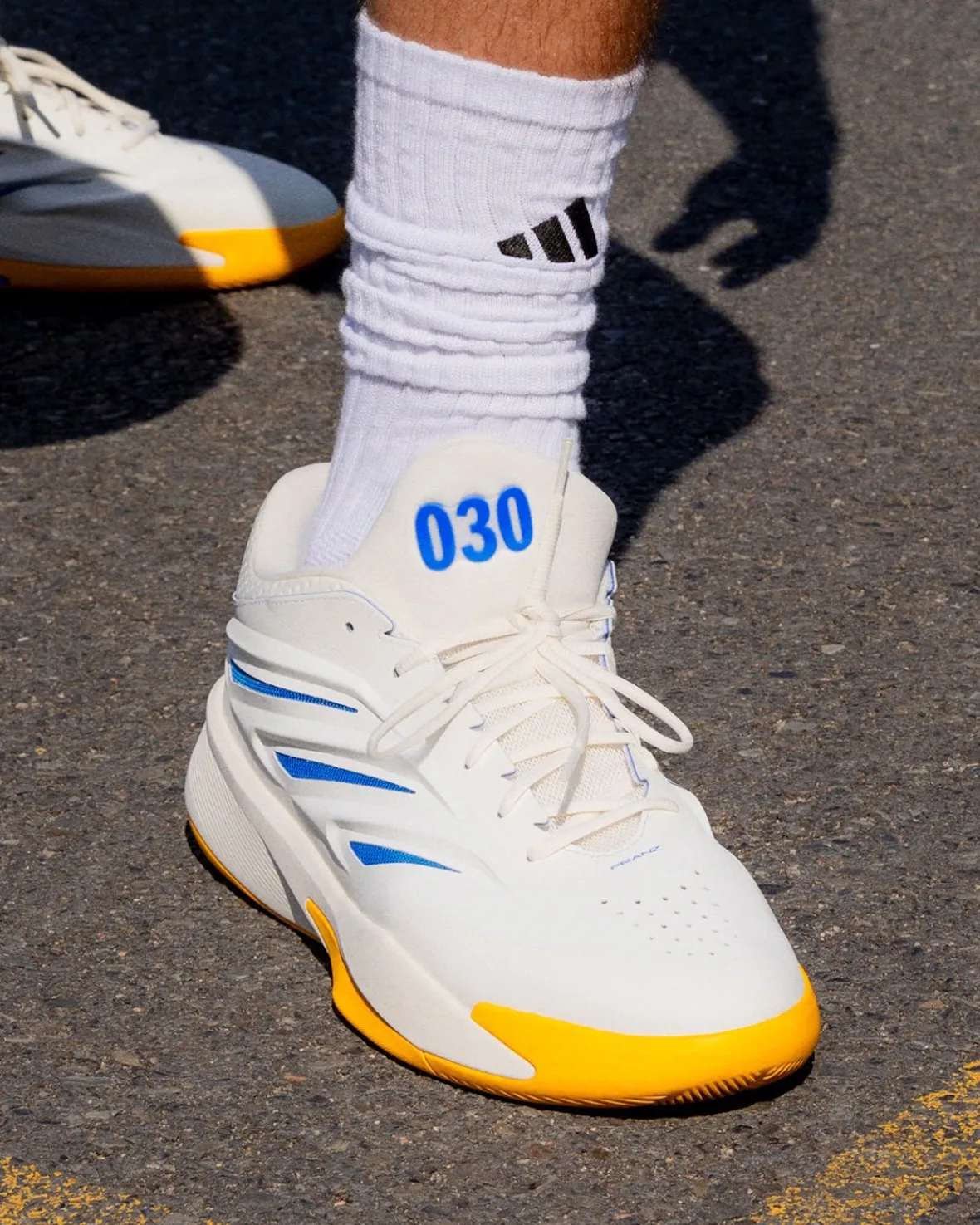 On-foot close-up of a white sneaker with blue 030 on the tongue, yellow sole and white sock, Wagner Brothers