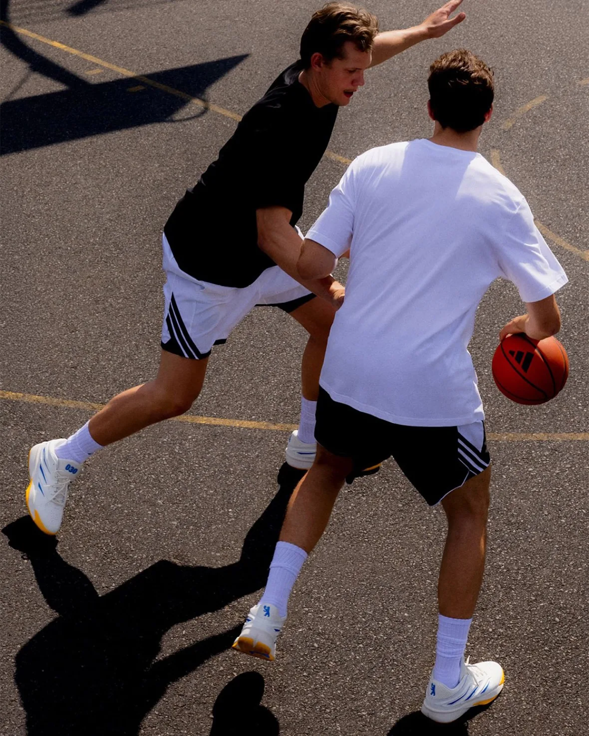 Overhead shot of two players in a one-on-one game on asphalt, wearing white and yellow sneakers, Wagner Brothers