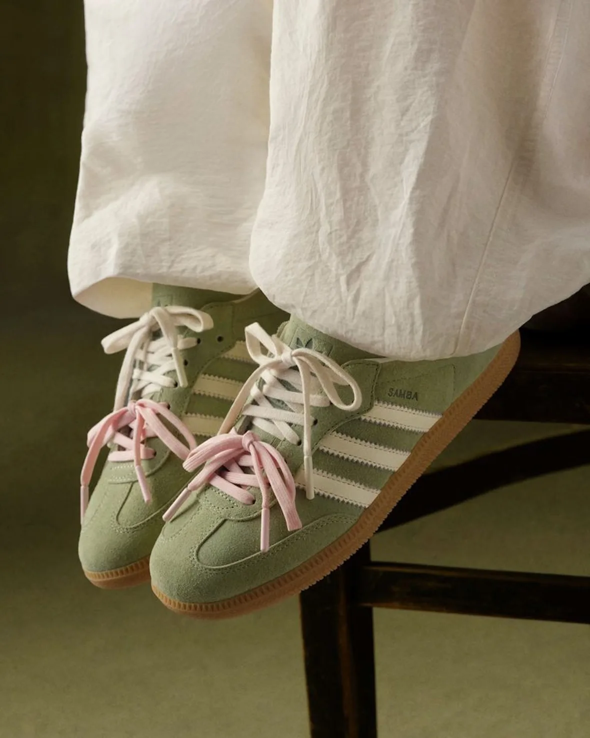 On-foot shot of green suede sneakers with pink and cream bows, cream trousers, wooden chair, Matcha