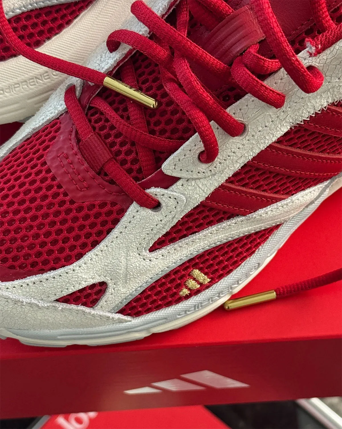 Close-up of red mesh, cracked white overlays, gold lace tip and gold three-stripe logo, x Tyrrell Winston