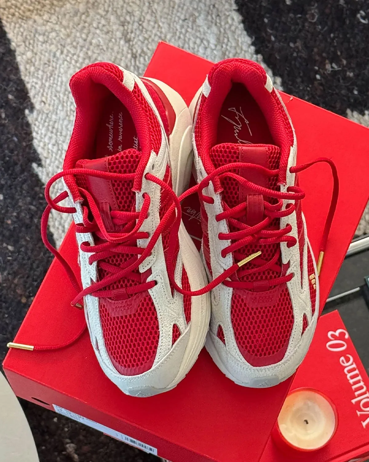 Top-down pair on a red box marked Volume 03, red laces with gold tips, candle nearby, x Tyrrell Winston