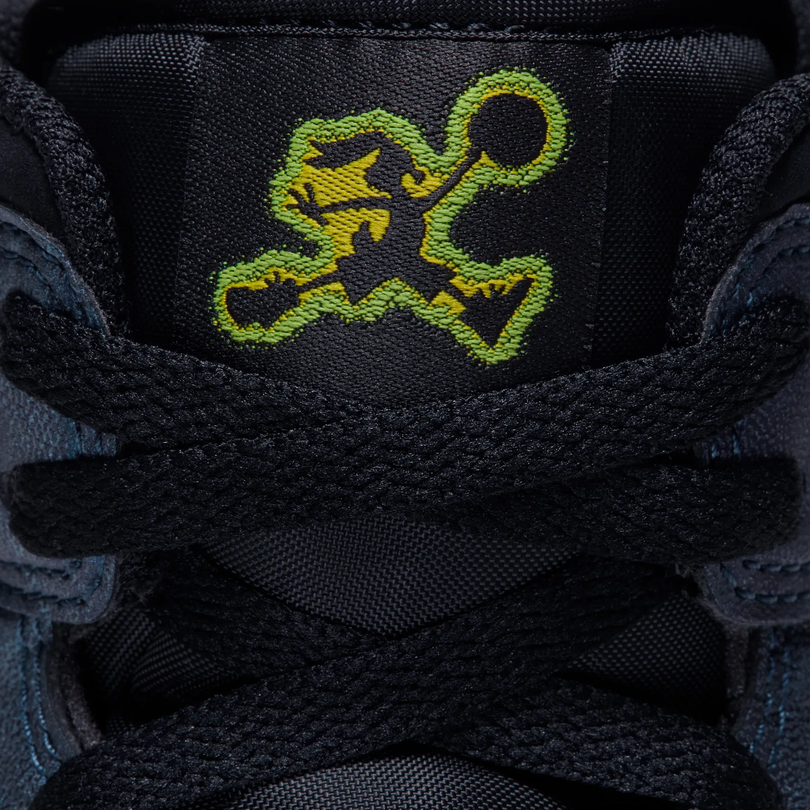 Close-up of tongue tag, green and yellow stitched cartoon jumpman on black fabric under crossed laces, Air Bugs