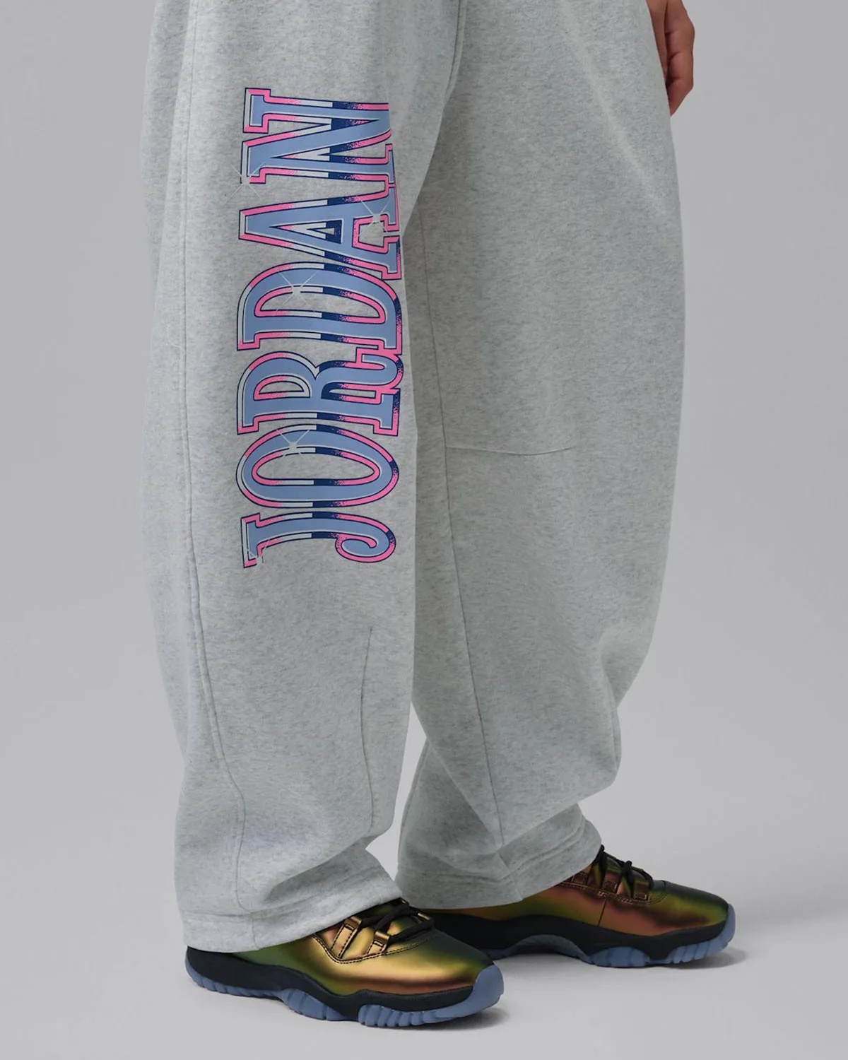 on-foot heel view, iridescent gold-green sneaker beside grey sweatpants with pink and blue Jordan wordmark, WMNS Space Jam
