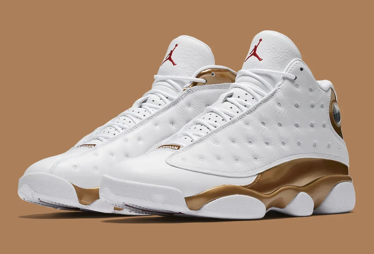 Pair of white sneakers with gold glossy panels and red Jumpman logos at an angle on brown background, Defining Moments Pack