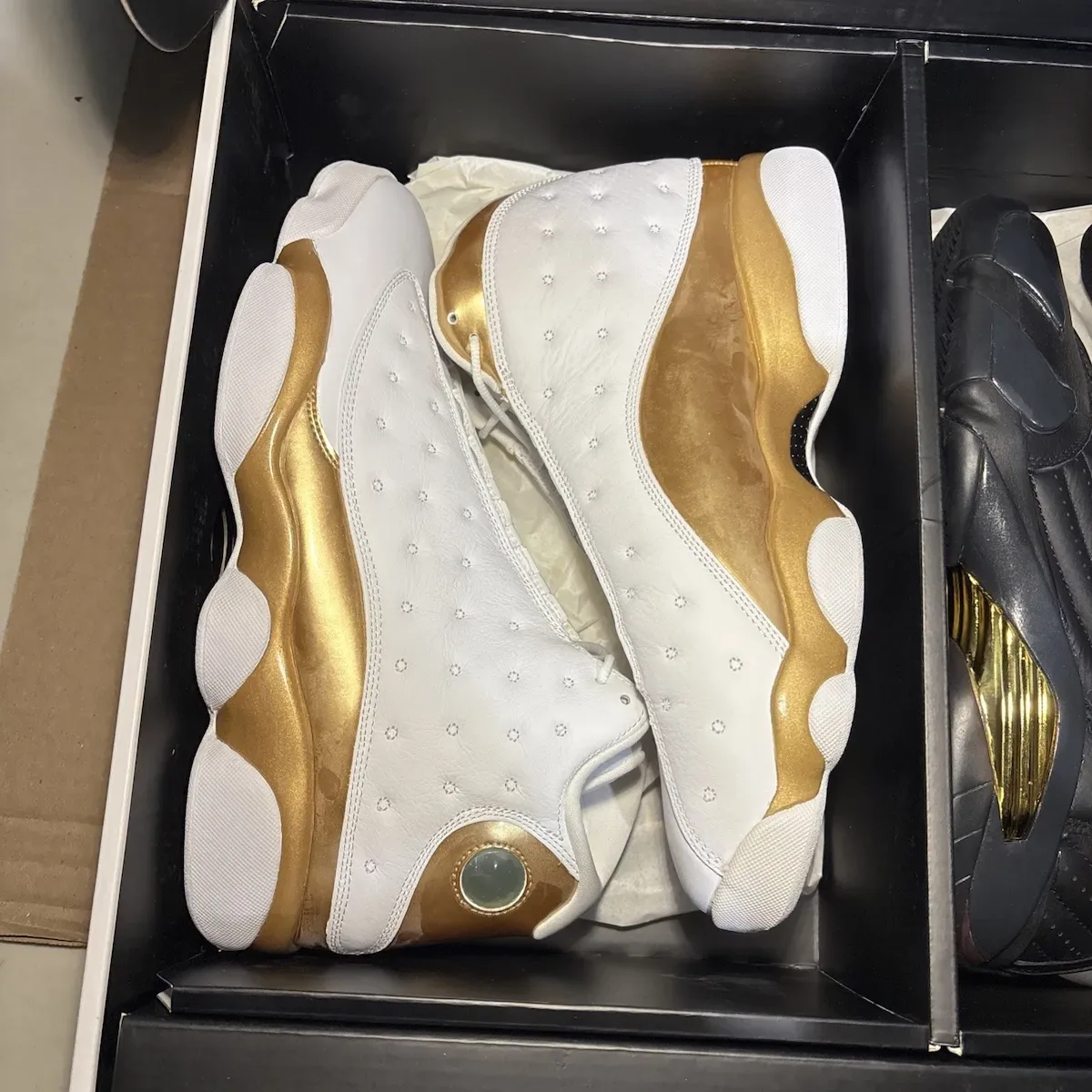 In-hand view in box of white sneakers with glossy gold panels and green circular heel badge, Defining Moments Pack