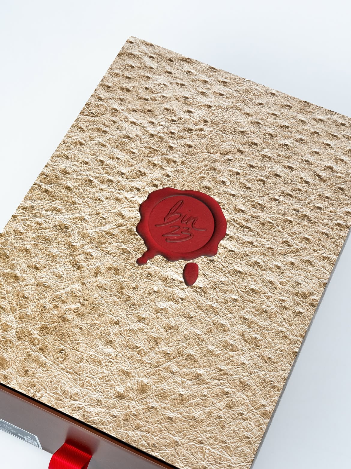 Box lid close-up, ostrich-textured tan with red bin23 wax seal and red ribbon pull, Bin23