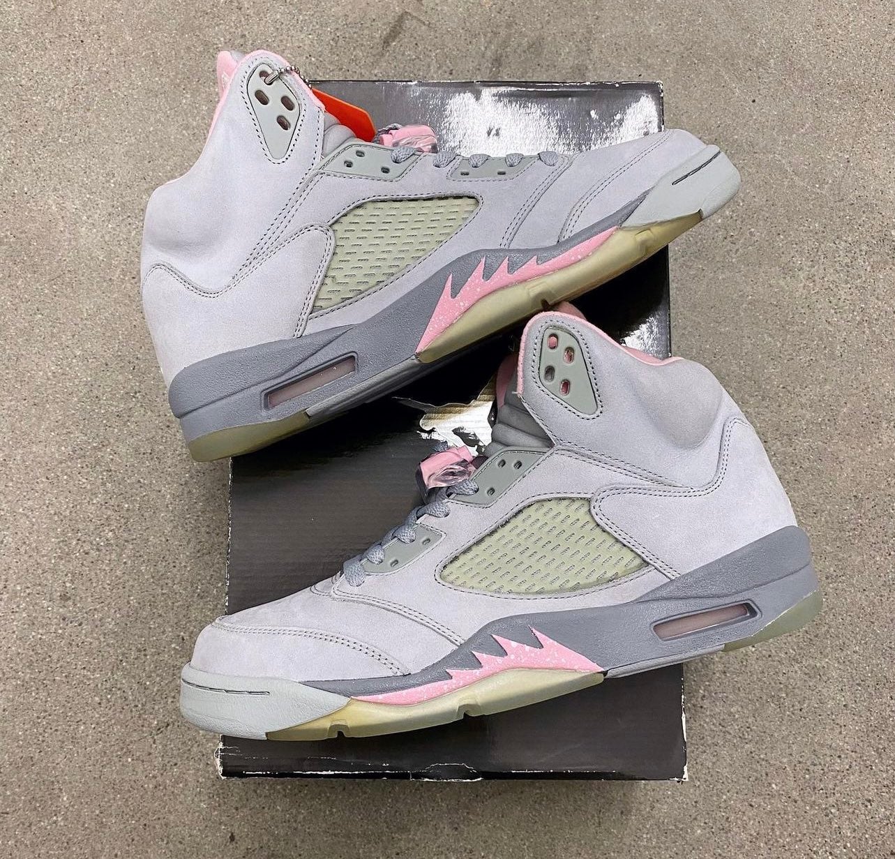 Pair stacked on a black box on concrete, grey suede with pink accents and orange tag, Shy Pink
