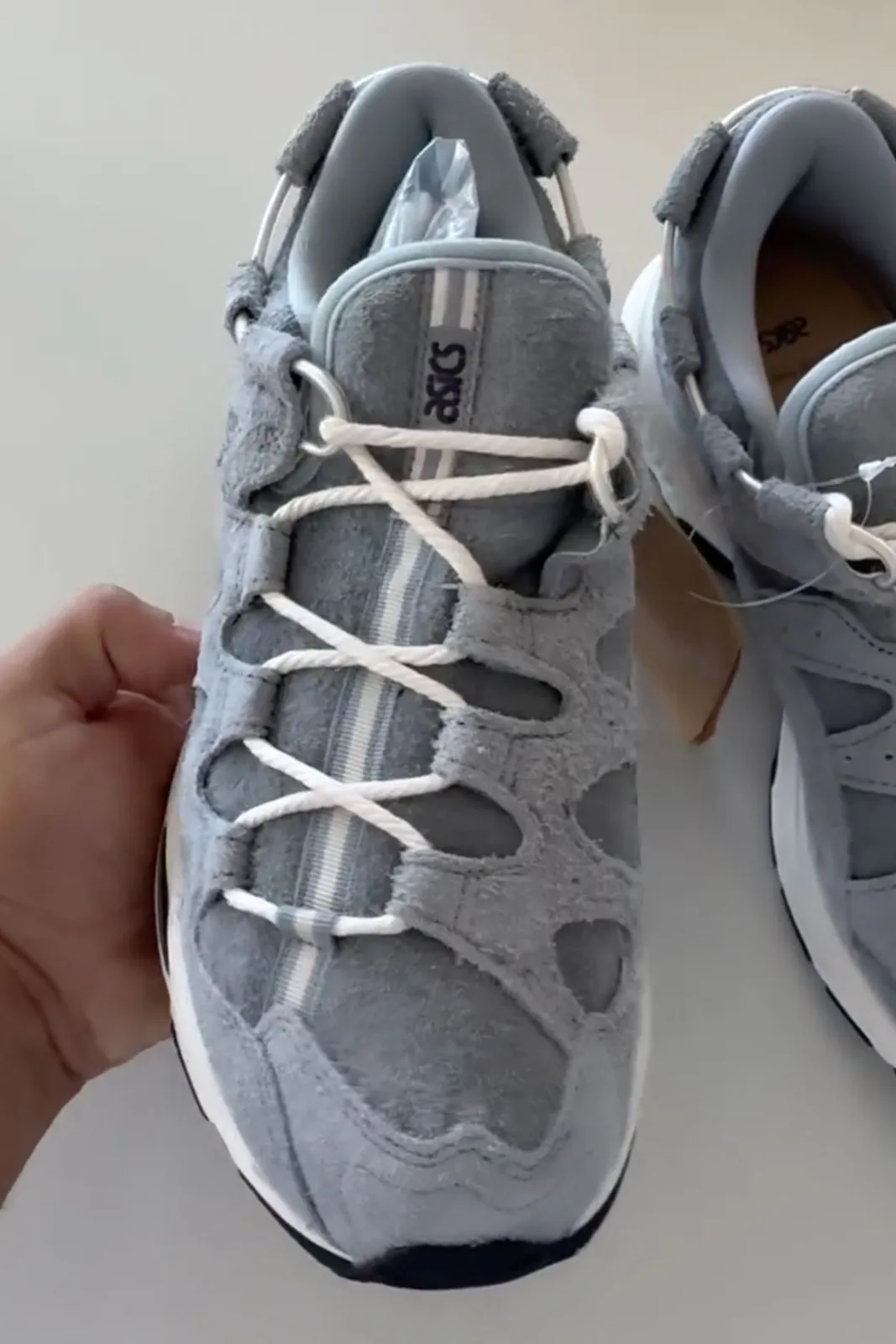 Top-down single shoe held in hand, blue-grey suede, white laces, ASICS tongue text, x Giorgio Armani