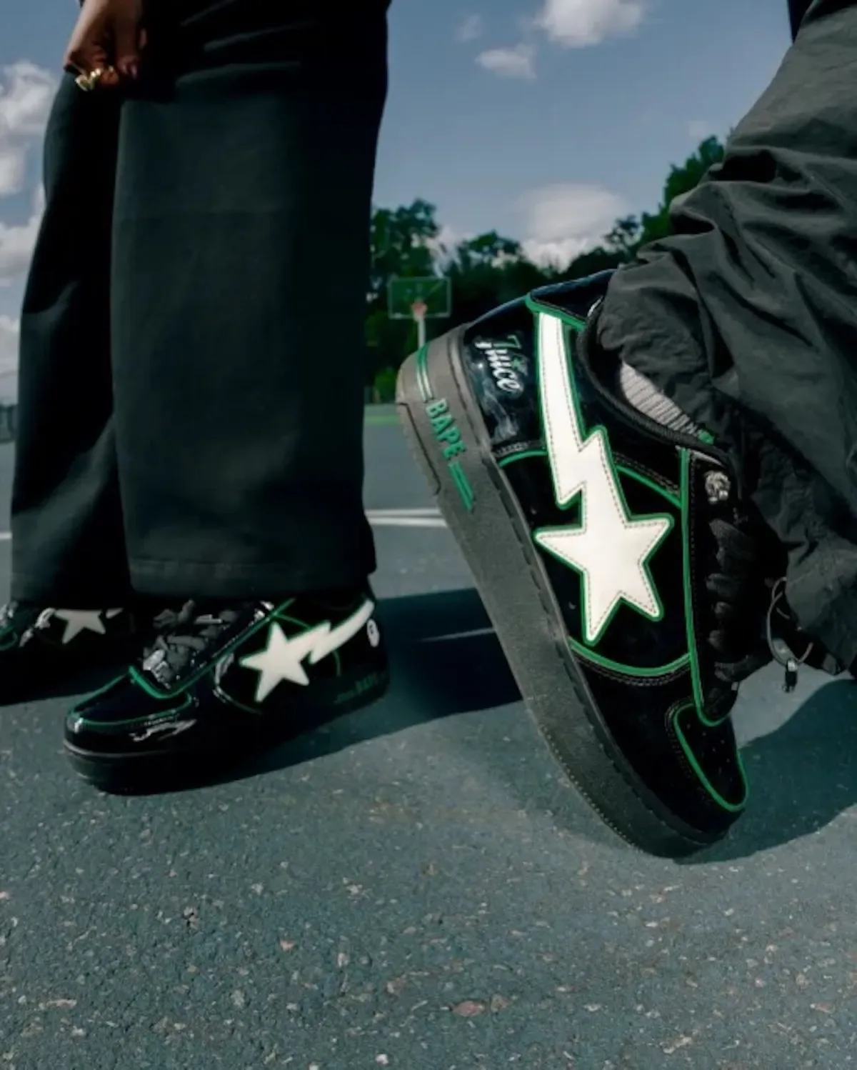On-foot lateral view of black patent sneakers with white star and green piping on an outdoor court, STA x 7UICE