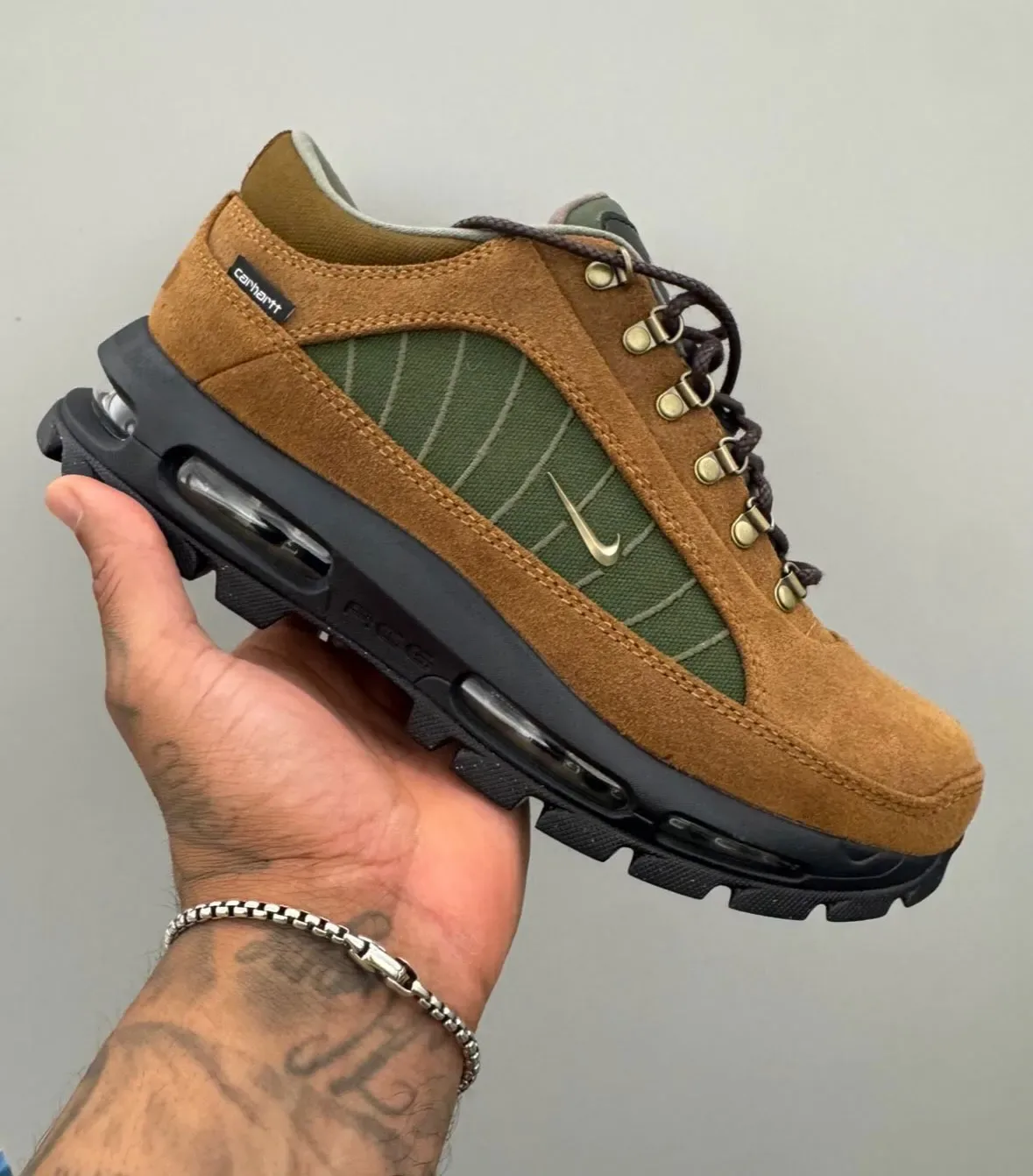 In-hand view of tan suede shoe with green panel, brass eyelets and tattooed hand, grey wall, Light British Tan