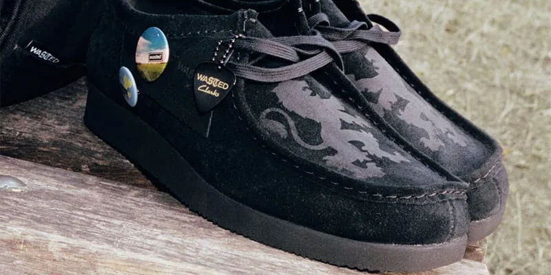 Close-up lateral view of black suede shoes with badges and a Wasted Clarks pick tag, x Wasted Paris