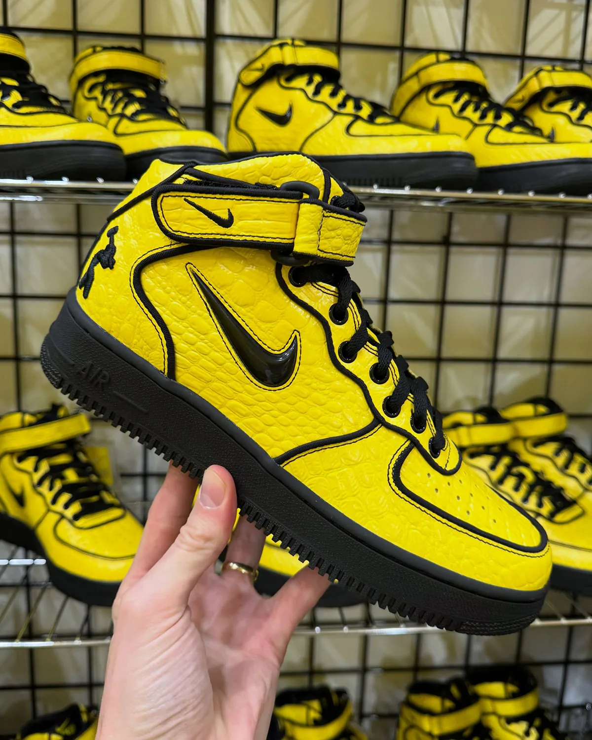 In-hand lateral view of yellow croc-embossed mid with black swoosh, shoes on wire shelves behind, Force 1 Mid