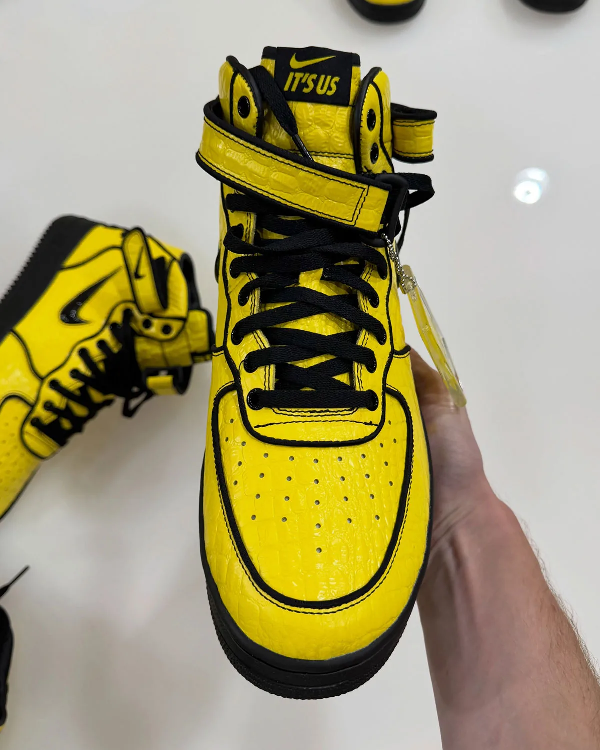 Top-down toe view in hand, yellow croc leather, black laces, "It's us" tongue label, Force 1 Mid