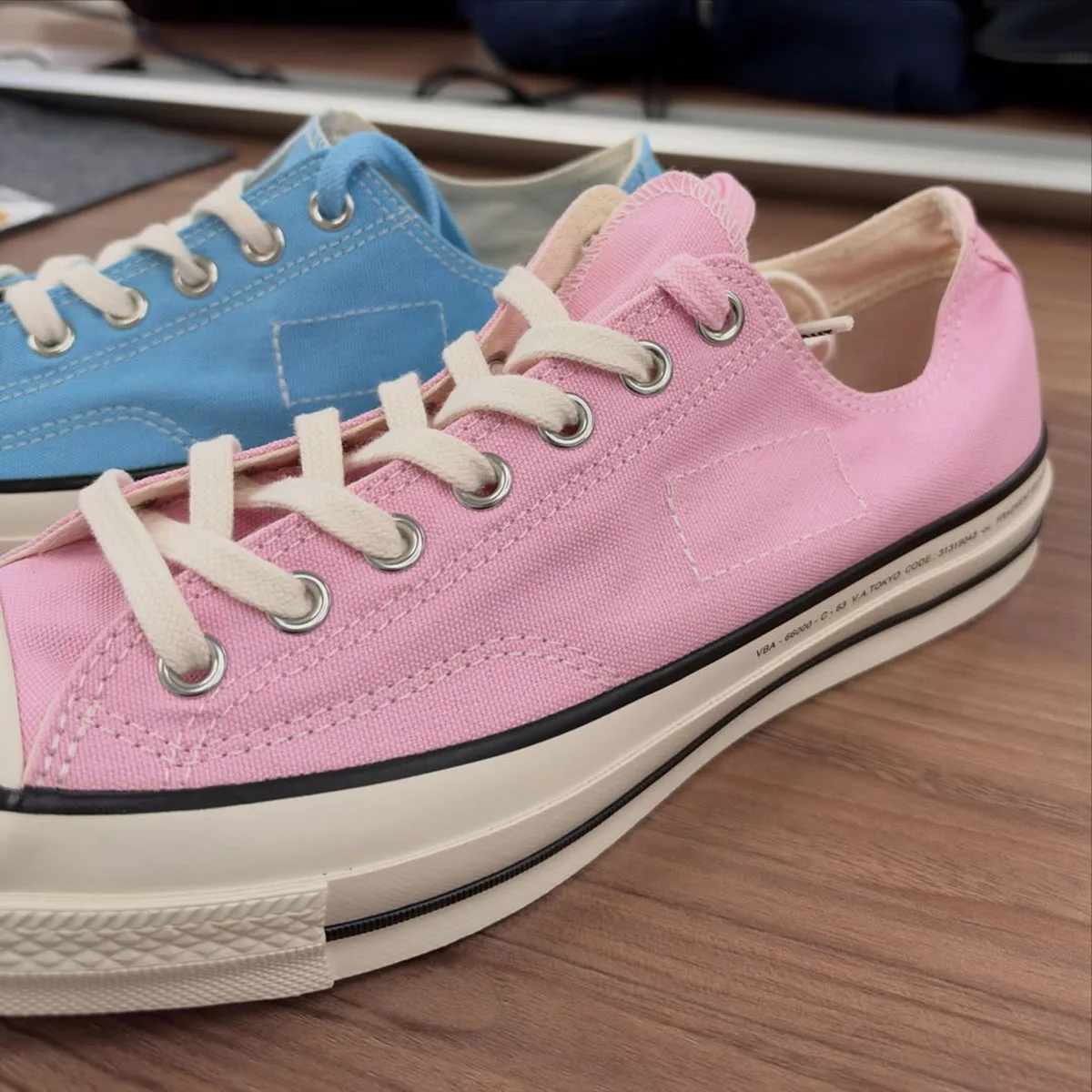 Lateral view of pink canvas low-top with cream laces, blue shoe behind, on wooden table, Chuck Taylor Low