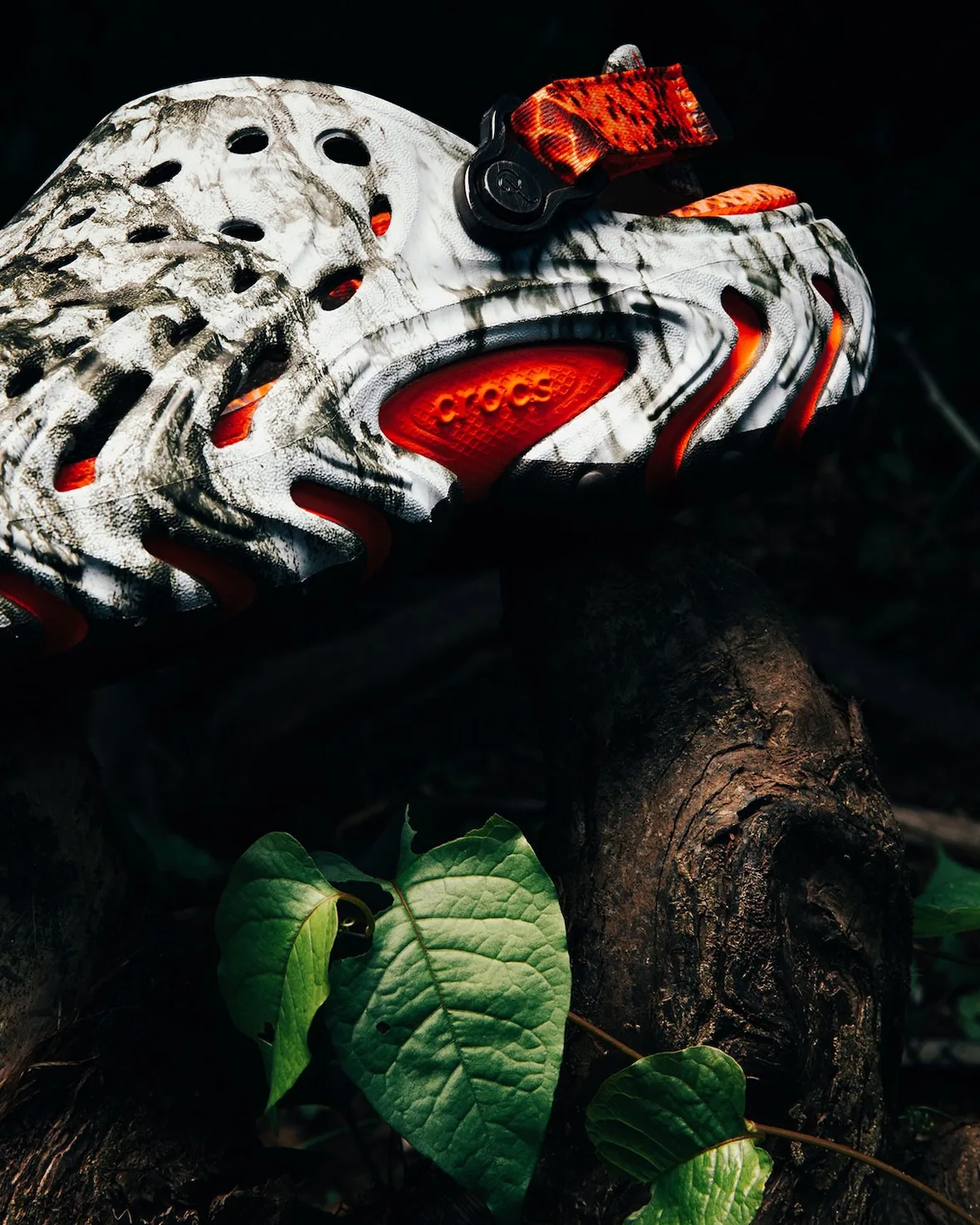 Lateral detail of marbled clog resting on root, orange crocs-embossed outsole pod, orange strap, green leaves, Watch Your