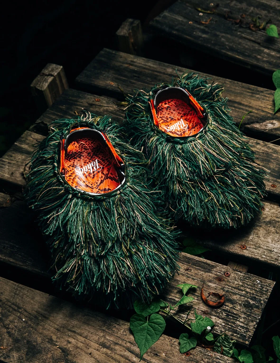 Pair of shaggy green fringe-covered clogs on wooden planks, orange snakeskin footbeds showing Politics script, Watch Your