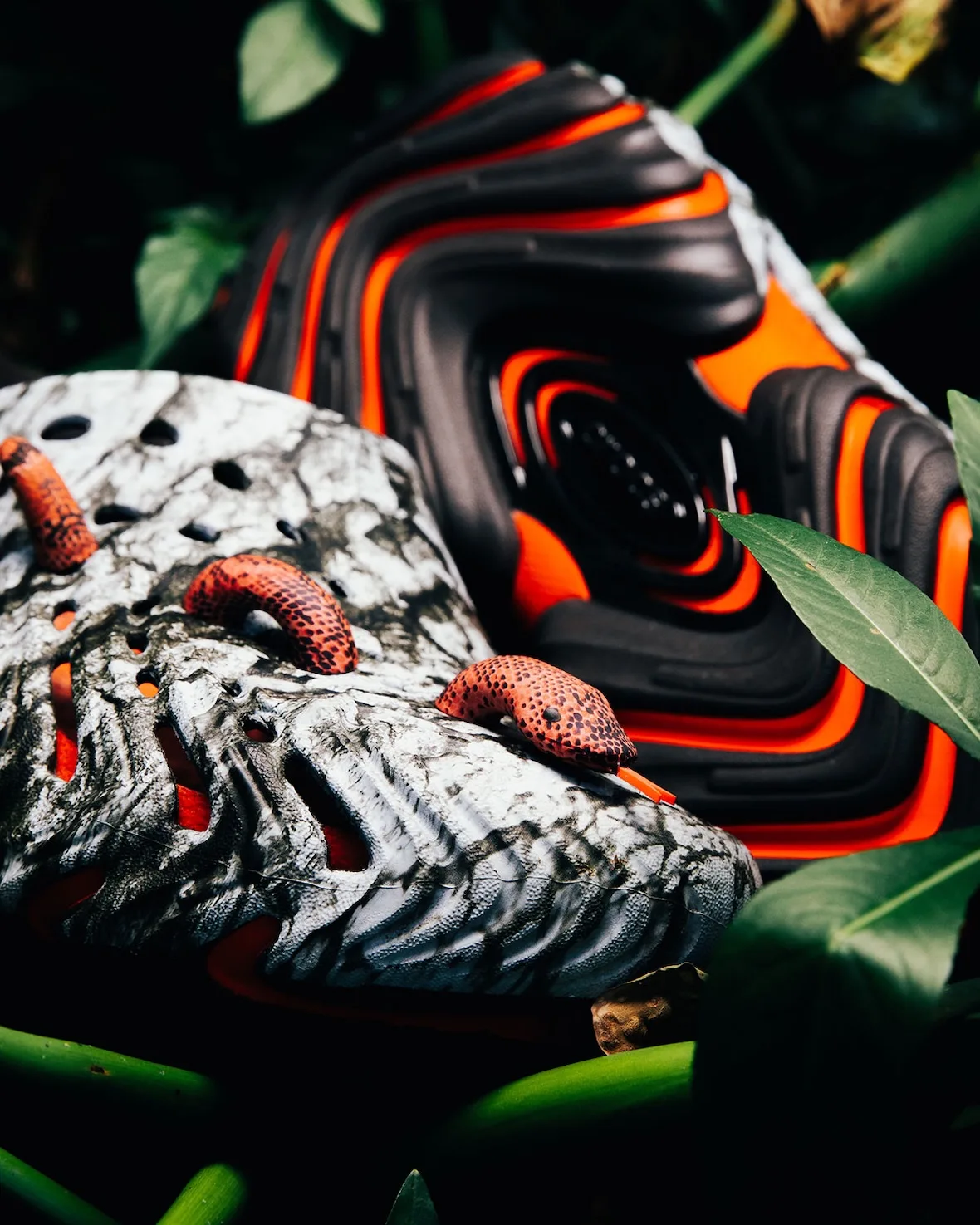 Close detail of orange snake charms on marbled clog toe, black orange tread behind, green leaves around, Watch Your Step