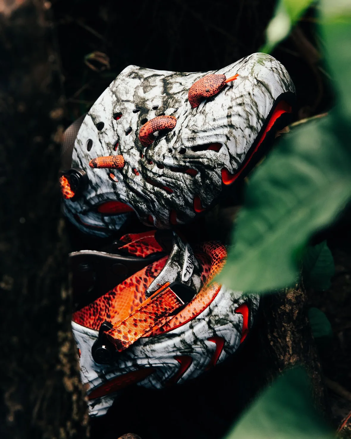 Detail of two marbled clogs among green leaves, orange snake charms on toe and orange snakeskin strap, Watch Your Step