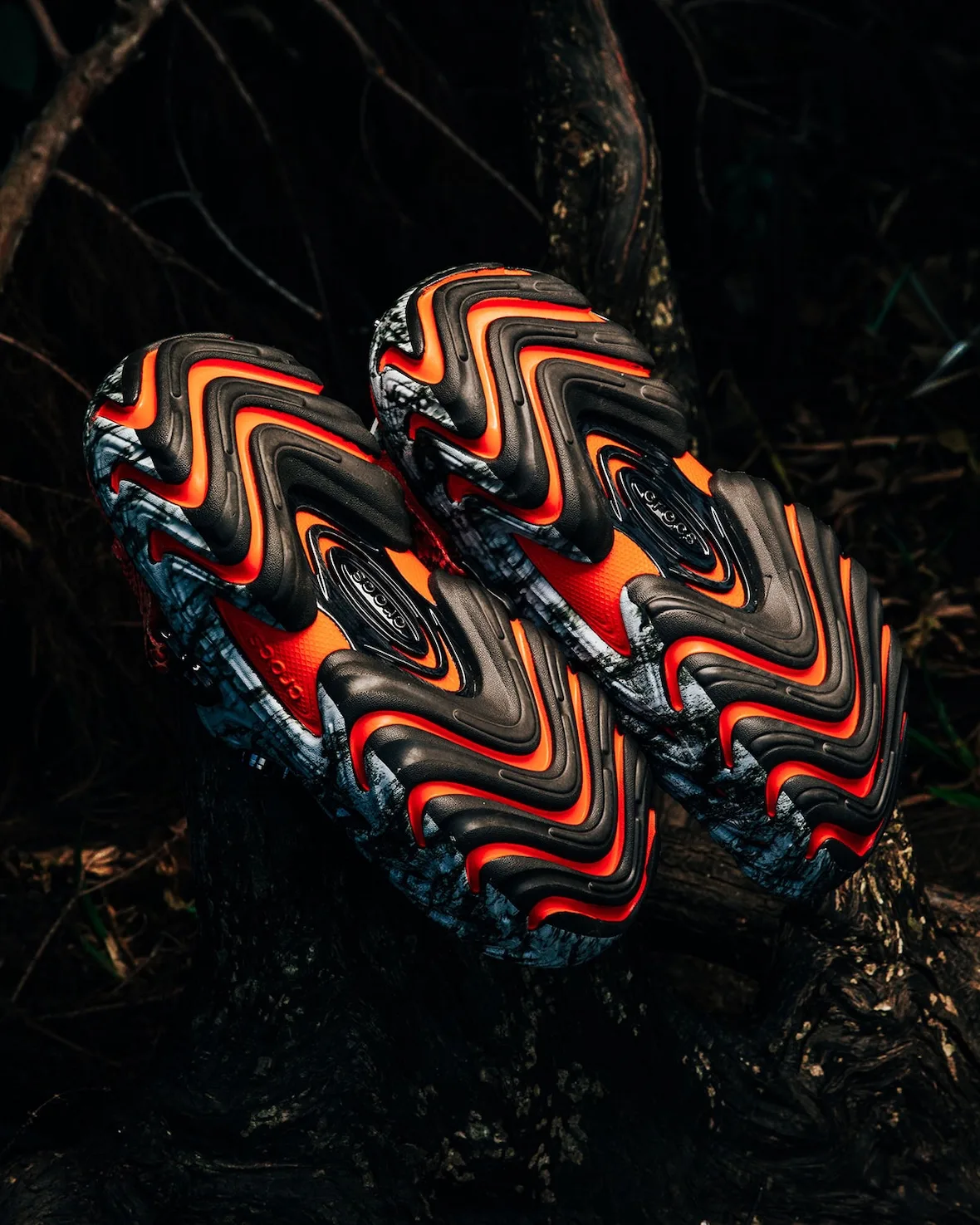 Outsole view of pair, black and orange wavy tread with marbled white-grey edges, on dark roots, Watch Your Step