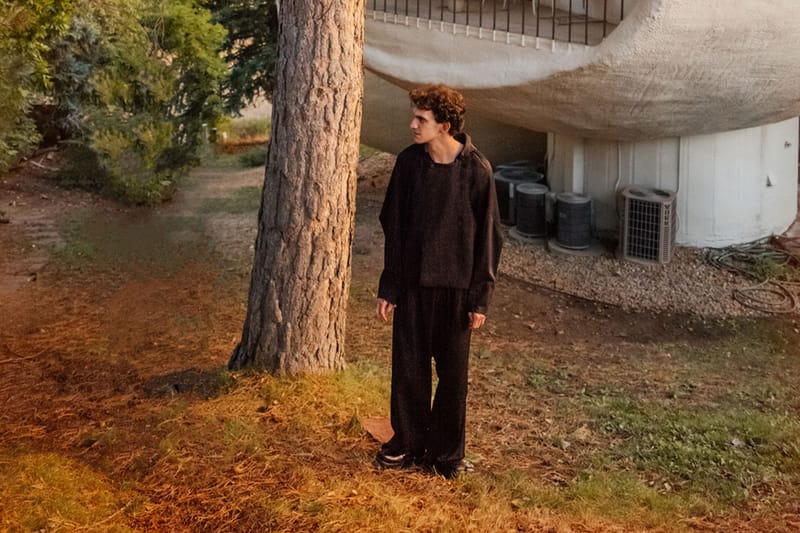 Model in dark outfit stands beside a tree in warm light, with a pale curved building behind, x Kiko Kostadinov