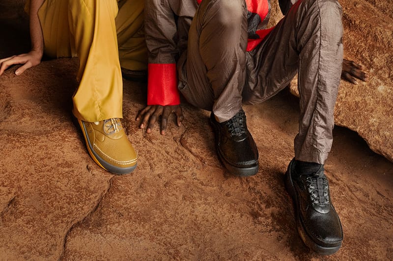Detail of mustard and black shoes on crouching people, with red cuffs and grey trousers on red rock, x Kiko Kostadinov