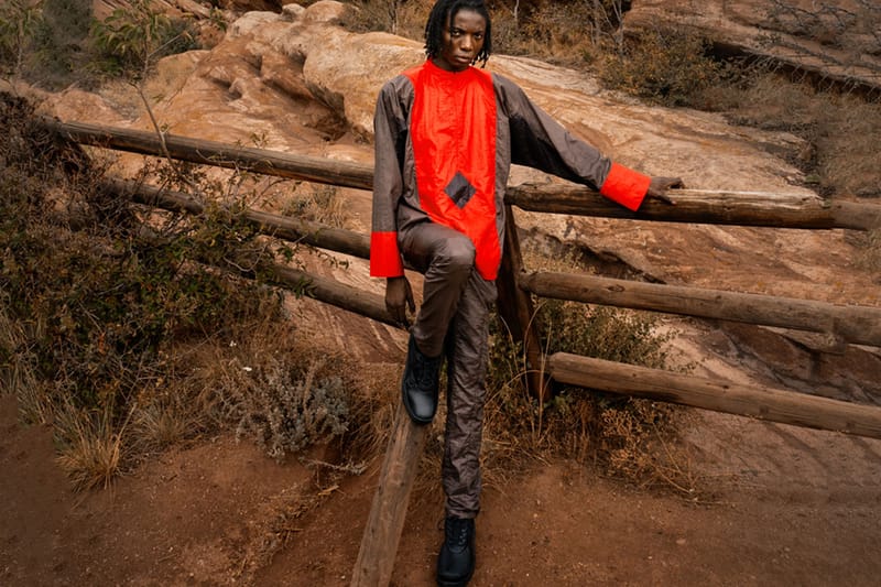 Model in red and grey jacket sits on a wooden fence in black boots, with red rocks behind, x Kiko Kostadinov