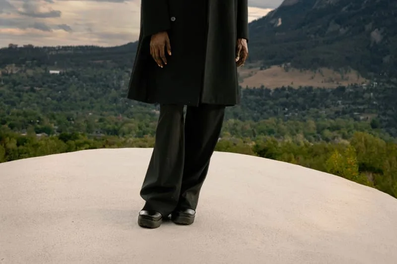On-foot view of black chunky shoes on a person in black coat and trousers on a pale dome above a valley, x Kiko Kostadinov