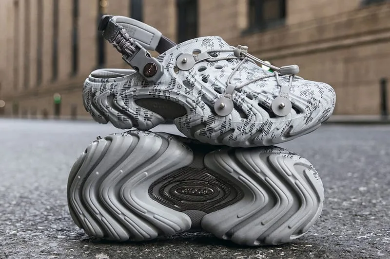 Pair on asphalt, grey digital-camo clog with toggle laces resting on outsole with Crocs logo, Echo Clog II