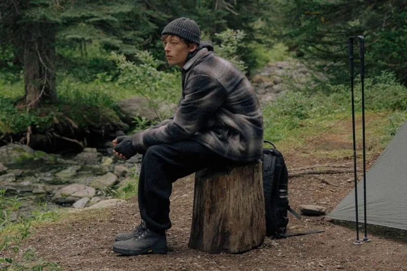 Model in beanie and plaid jacket seated on a stump, forest campsite, tent, poles, Black