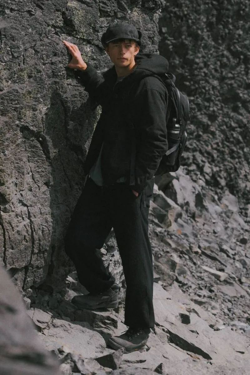 Model in black jacket and cap leaning on a rock face, backpack, dark boots, Black