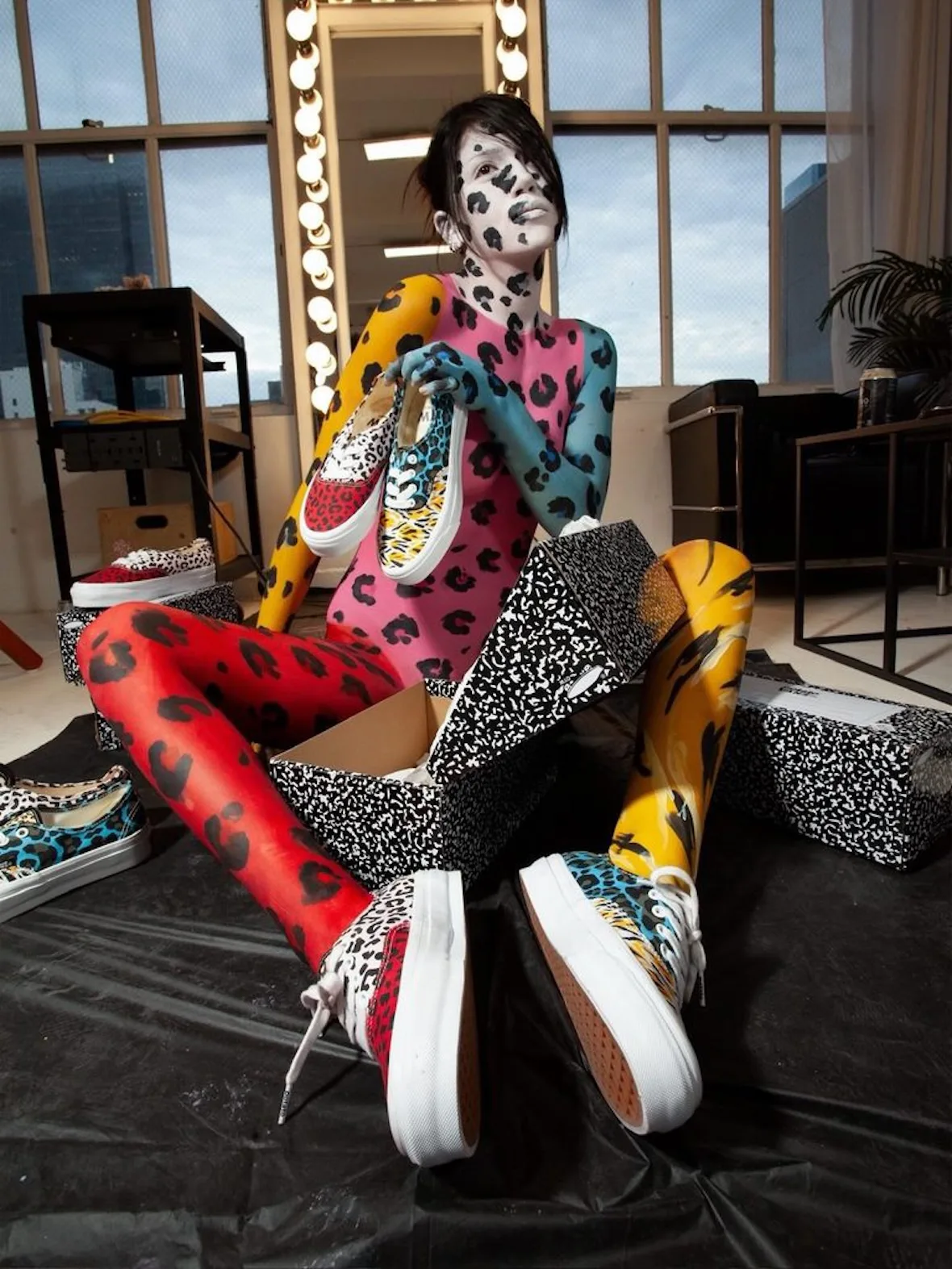 Body-painted model seated among speckled shoe boxes, holding and wearing red, cream and teal leopard-print sneakers, Cheetah