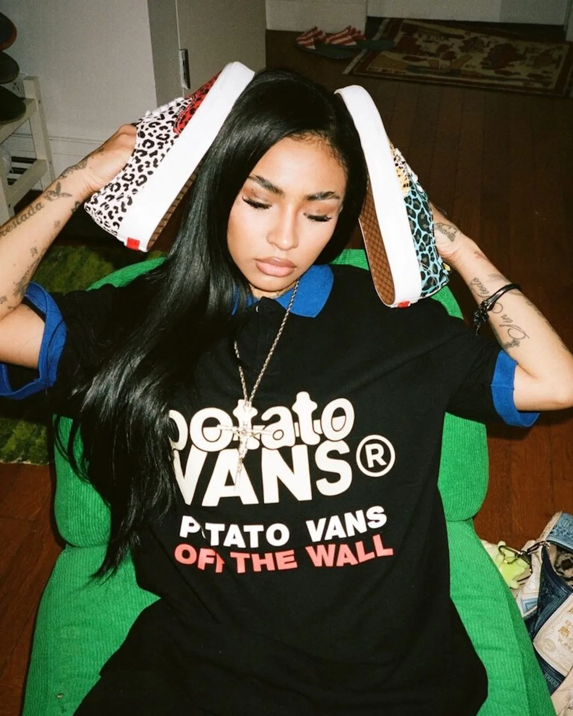 Woman in black Potato Vans tee holding multicolour leopard sneakers above her head, green chair behind, Cheetah Print