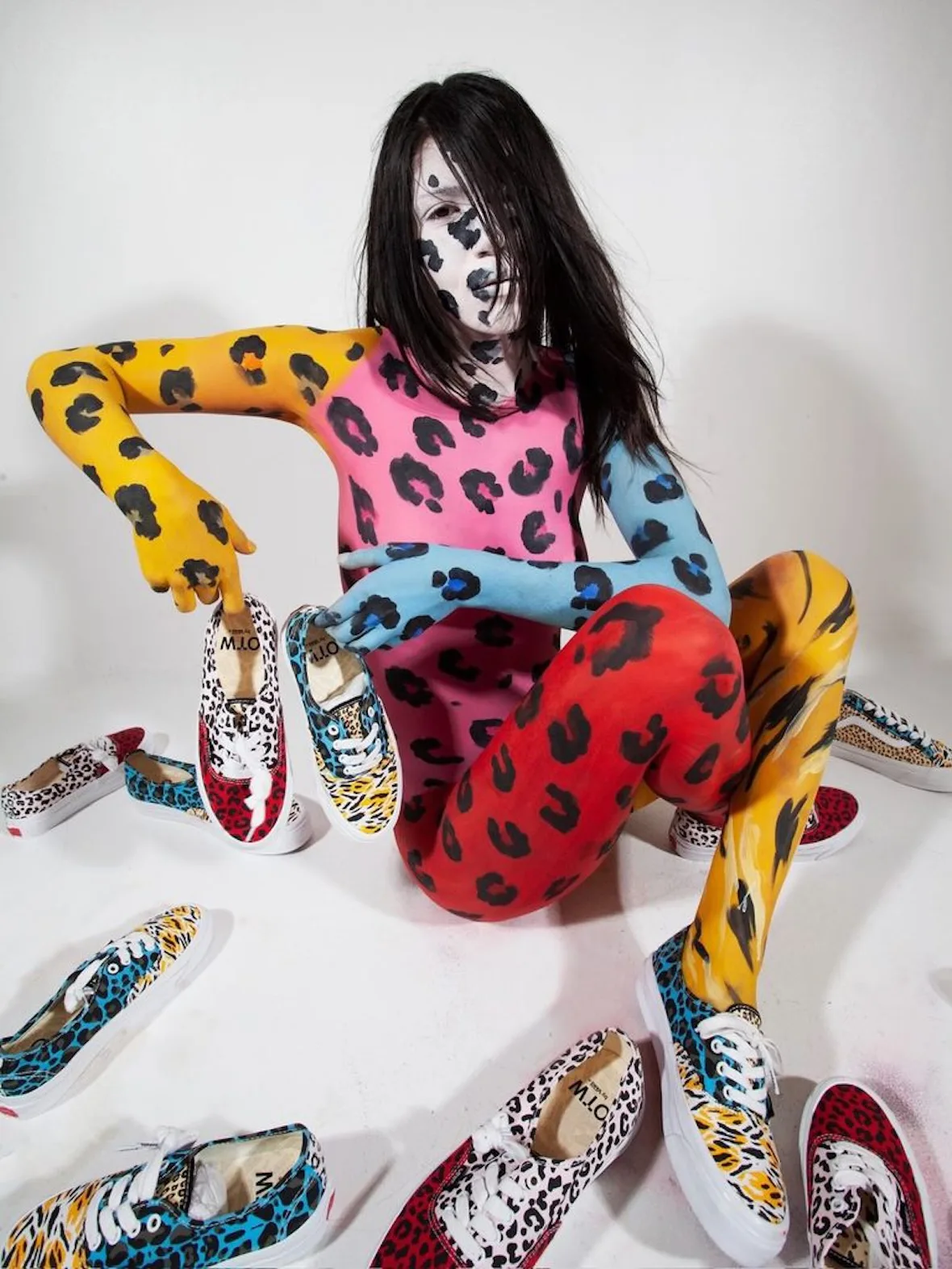 Body-painted model on white floor holding leopard-print sneakers, more red, teal and cream pairs scattered around, Cheetah
