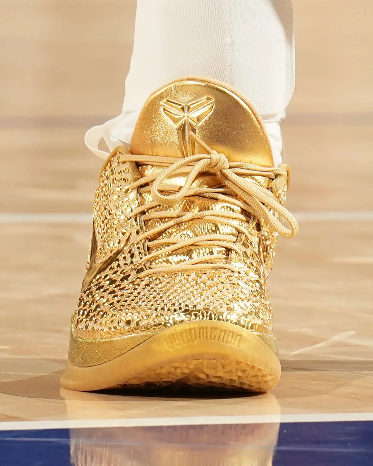 Heel-on view on court of a gold shoe with embossed logo on the heel tab, tan laces and gold outsole, Ring Night