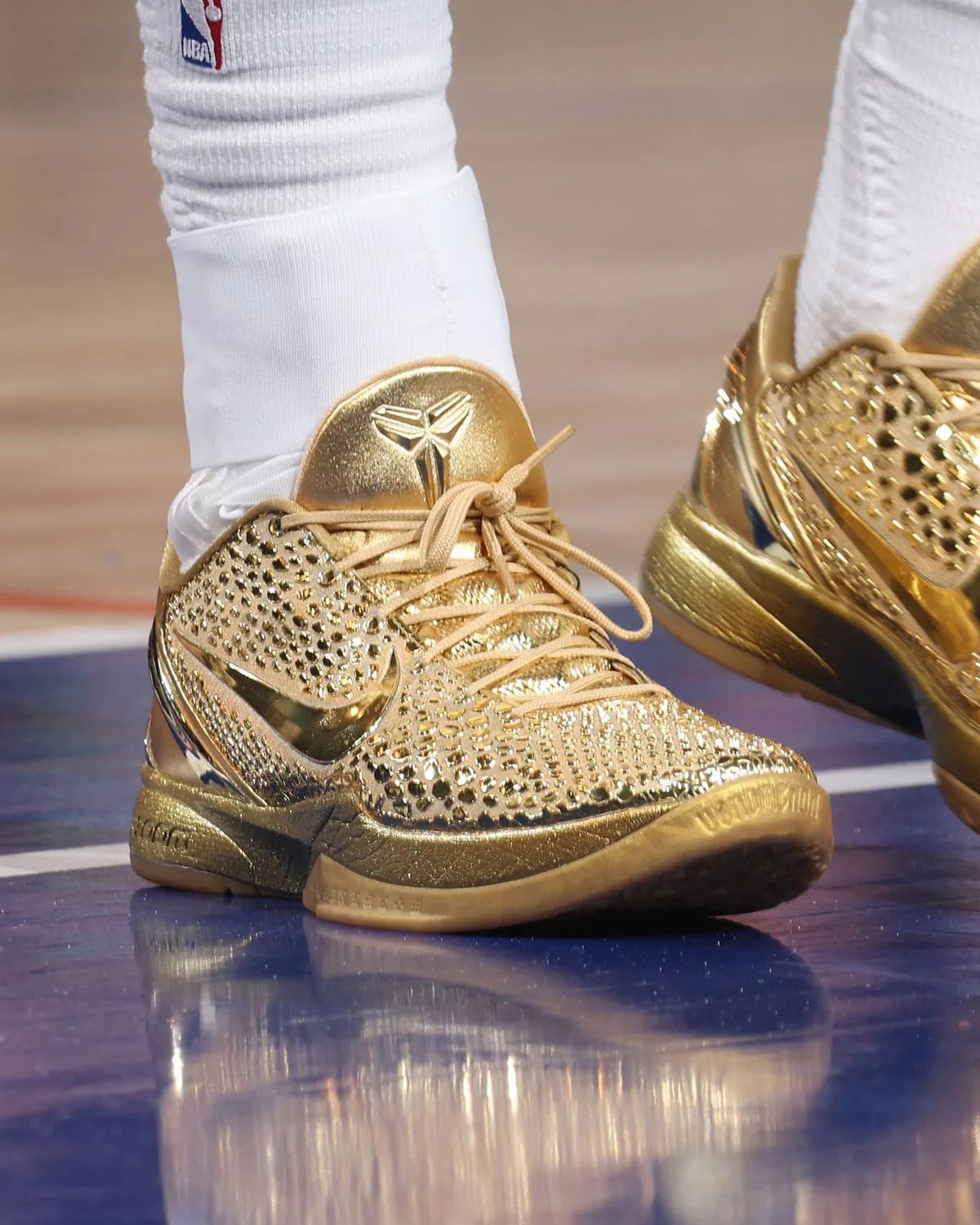 On-foot lateral view of gold shoe on a blue and wood court, gold heel logo, tan laces and reflective floor, Ring Night