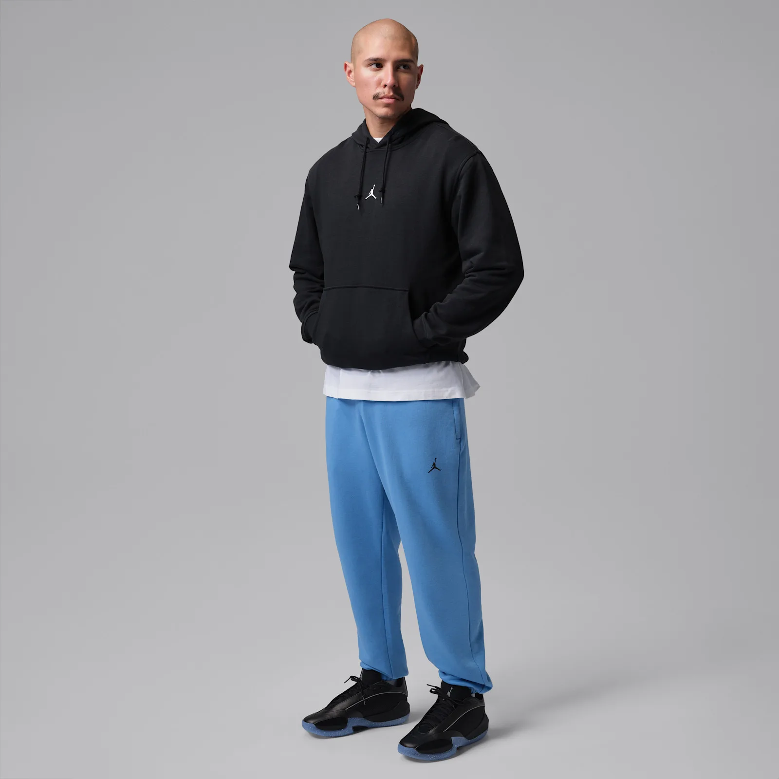 On-foot full-body shot, black sneakers with blue sweatpants and a black hoodie, grey backdrop, Black/Metallic Silver