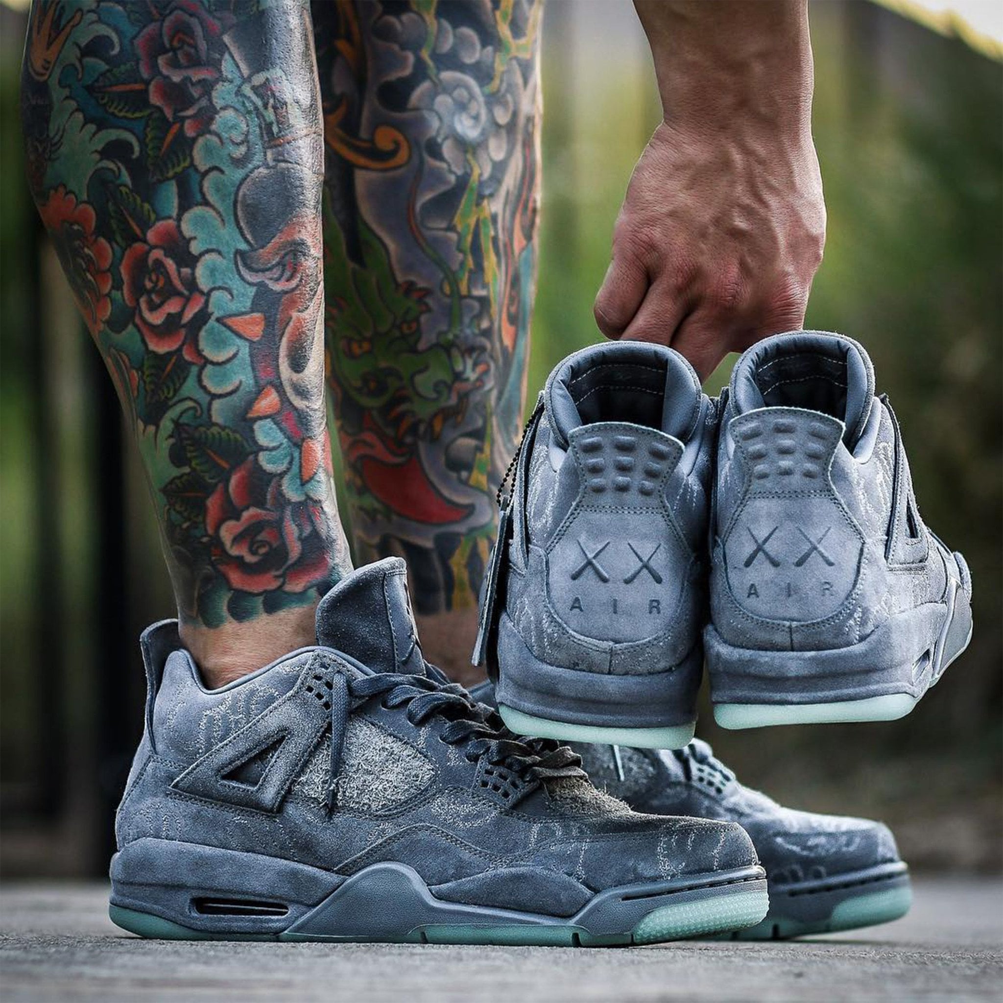 On-foot shot of grey suede sneakers, hand holding pair by heels with XX AIR logo, tattooed legs, Dark Grey