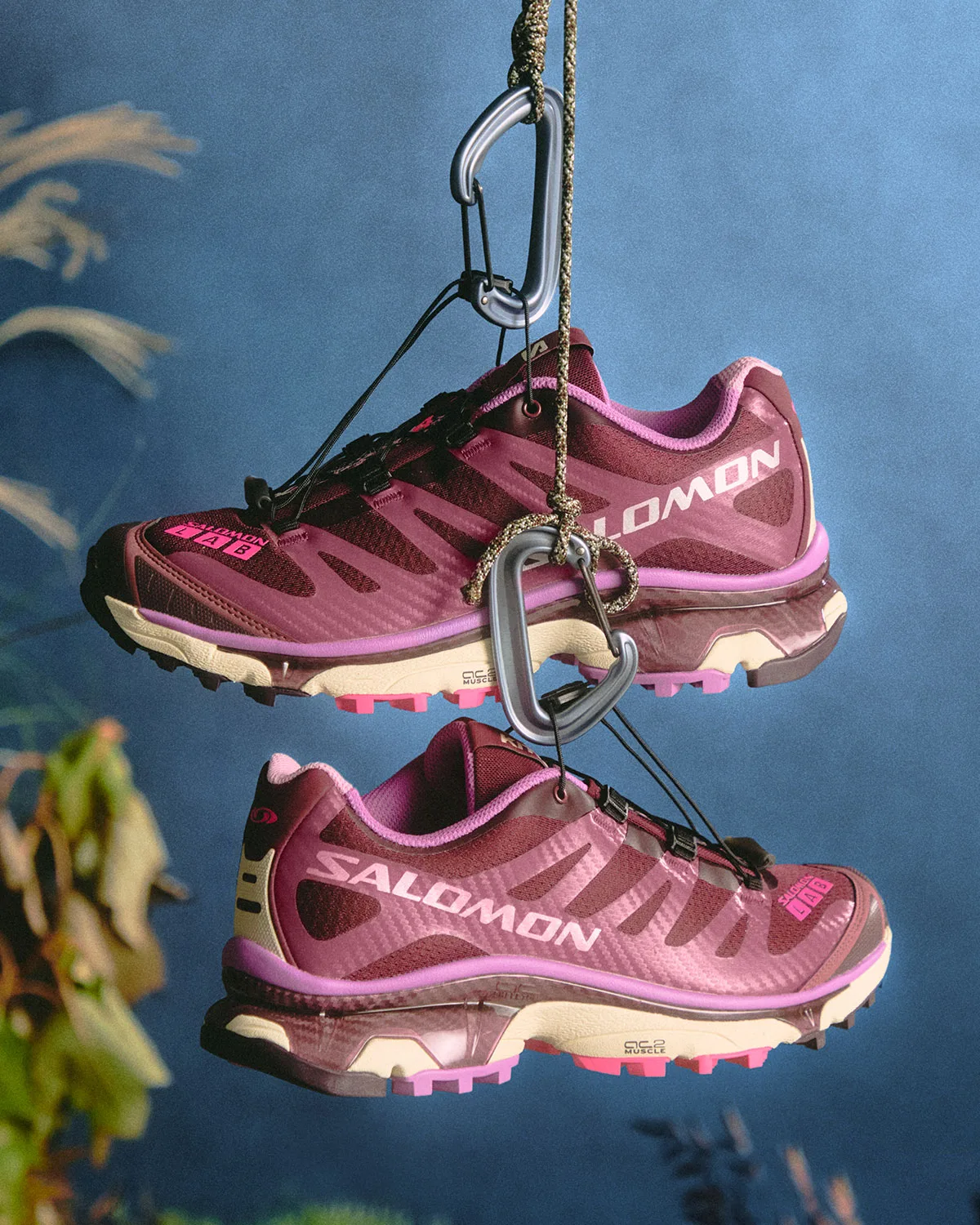 Pair of burgundy shoes with purple trim and pink lugs hanging from carabiners, blue backdrop, Winter 2026 Collection