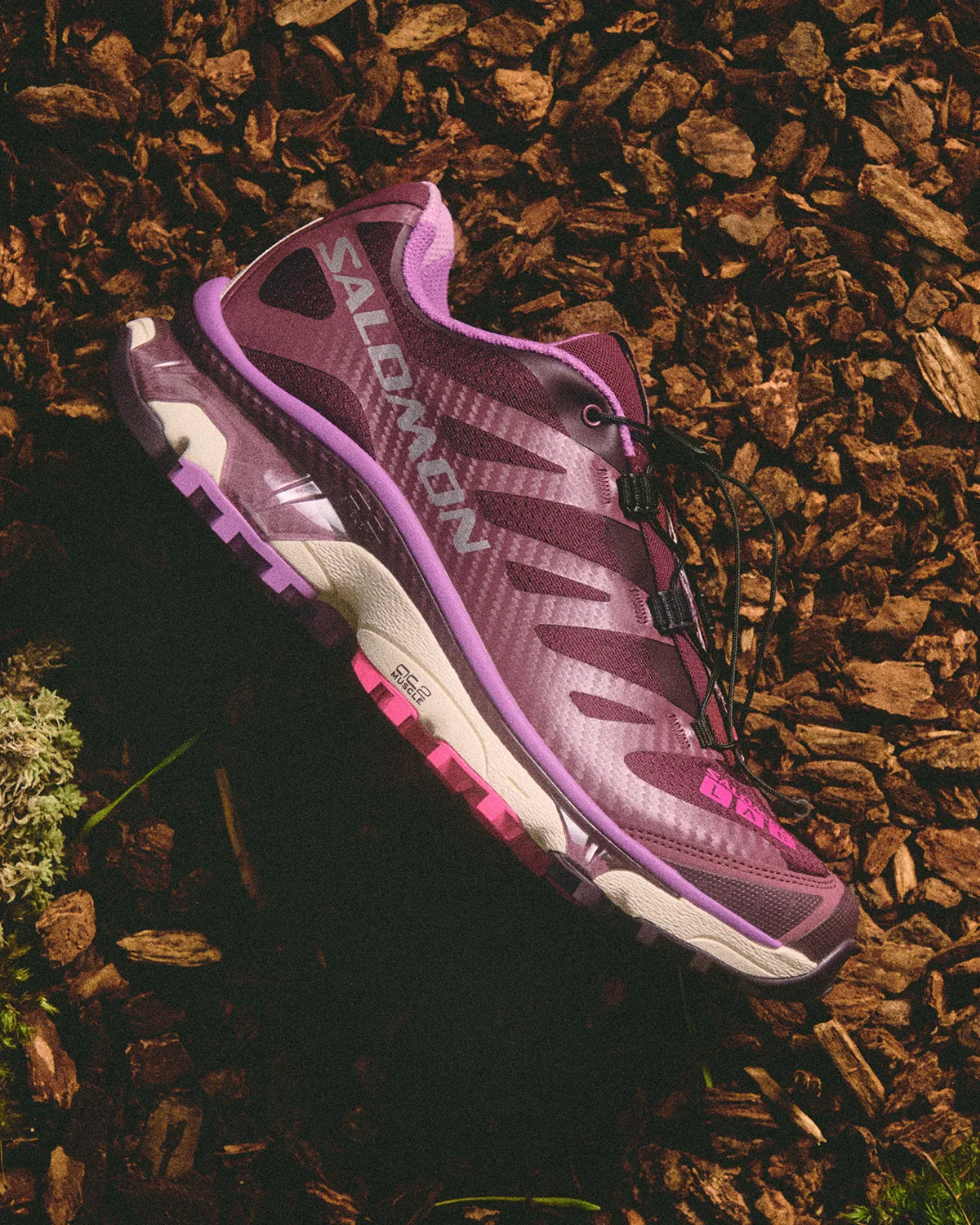 Lateral view of a burgundy shoe with purple trim and pink heel lugs on bark mulch, Winter 2026 Collection