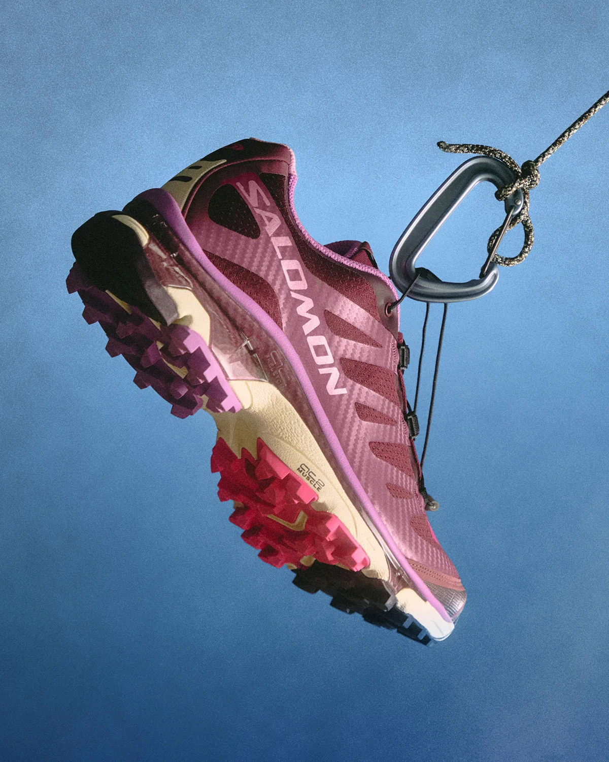 Lateral view of a burgundy and purple shoe hanging from a carabiner, pink lugs, blue backdrop, Winter 2026 Collection