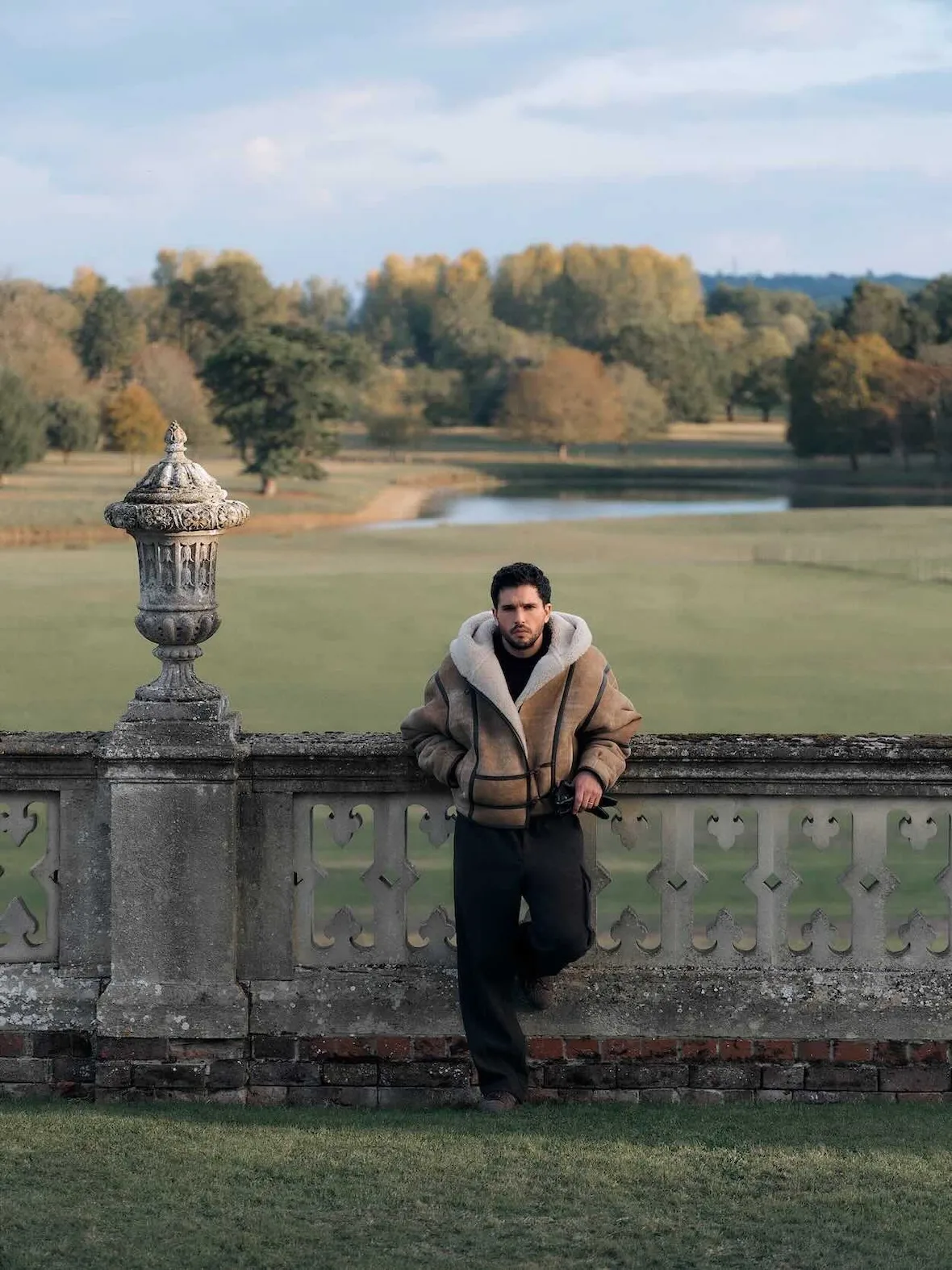 On-model leaning on stone balustrade, shearling hooded tan jacket, park lake and autumn trees behind, Winter 2026 Collection