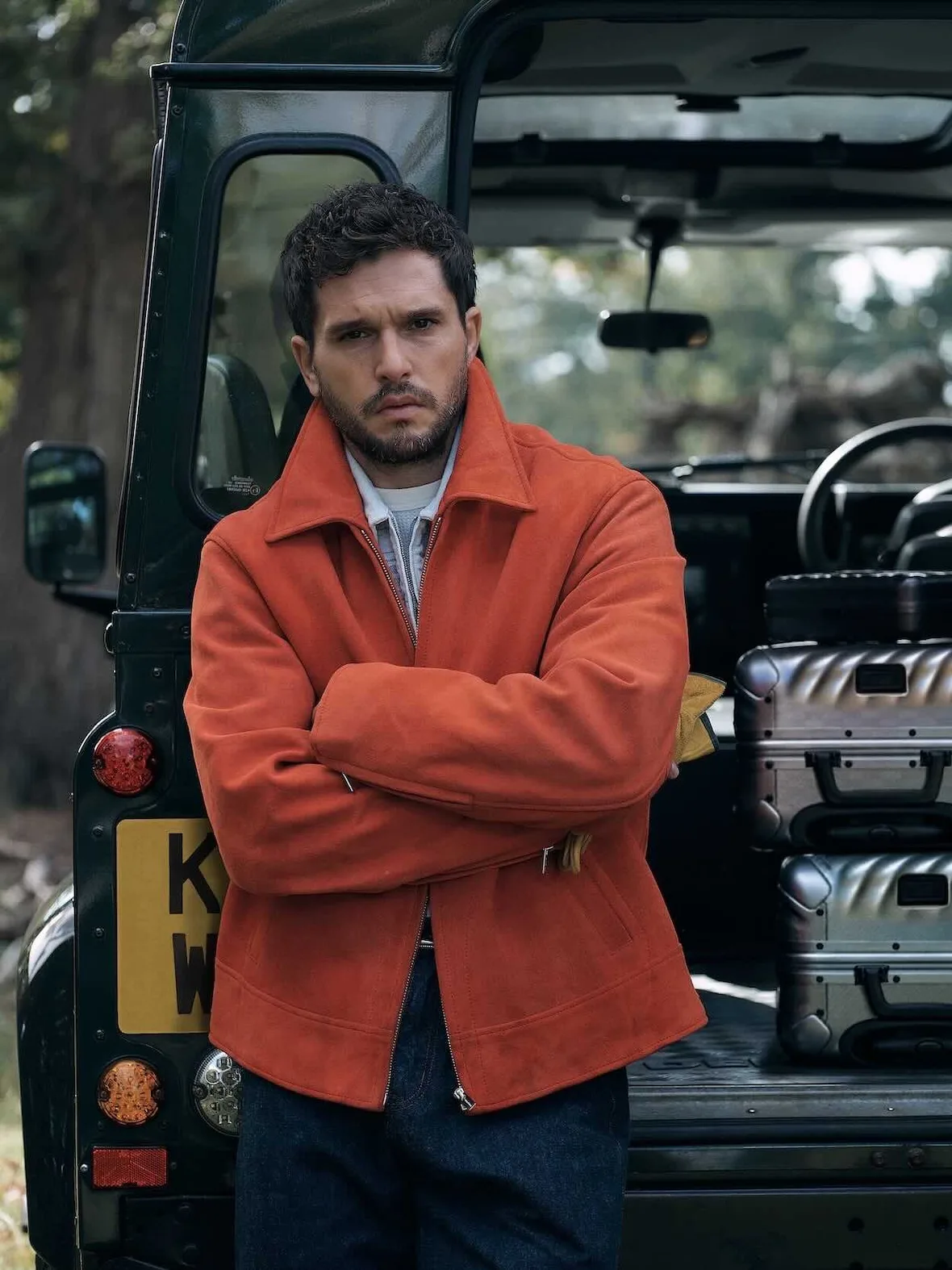 On-model, orange suede jacket, arms crossed, leaning on dark green 4x4 with yellow plate and silver luggage, Winter 2026