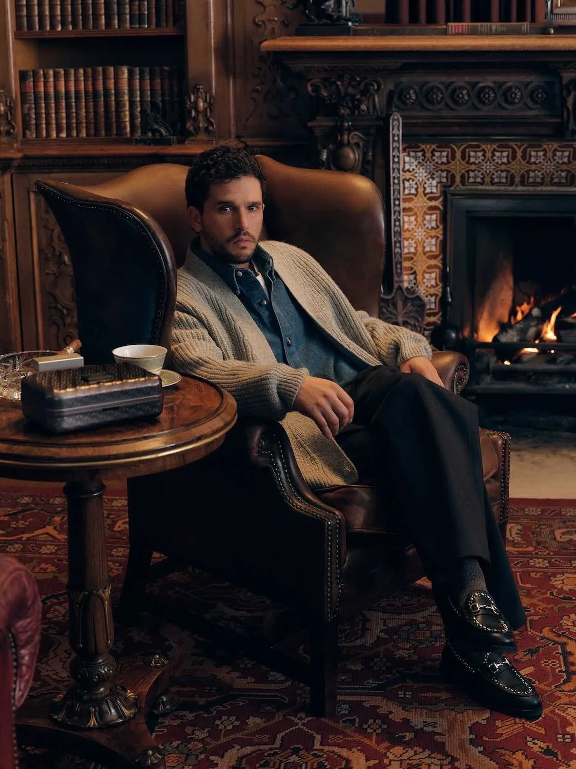 On-model seated by lit fireplace in leather armchair, beige cardigan, black horsebit loafers, tiled surround, Winter 2026