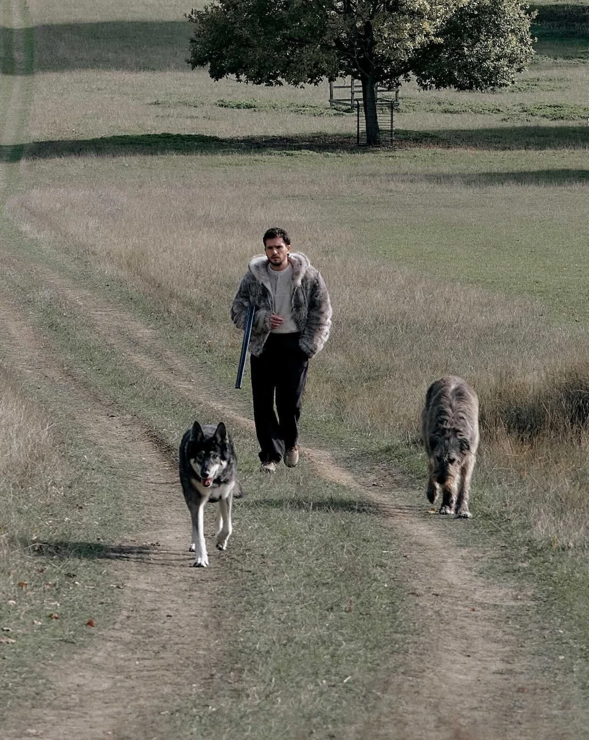 Wide shot of man in fur hooded jacket carrying shotgun on dirt track, flanked by two dogs in field, Winter 2026 Collection