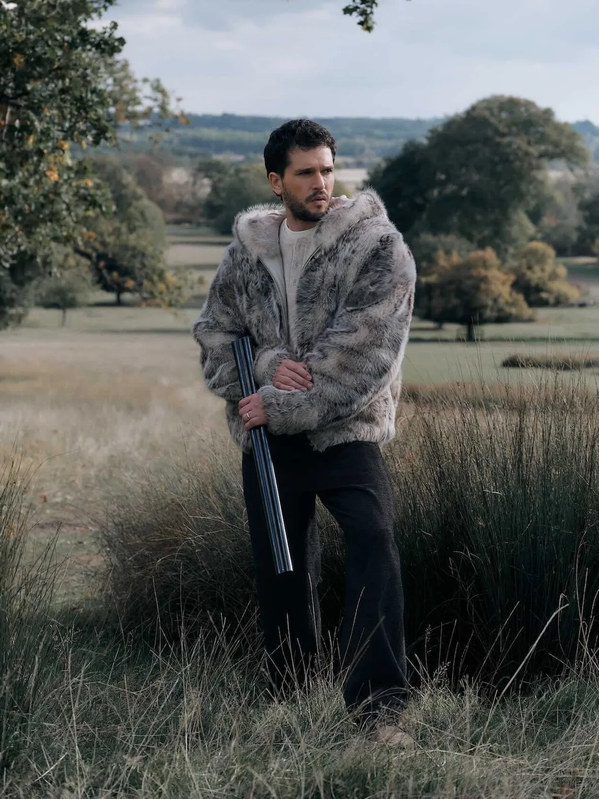 On-model standing in tall grass, grey fur hooded jacket, black trousers, holding broken shotgun, trees behind, Winter 2026