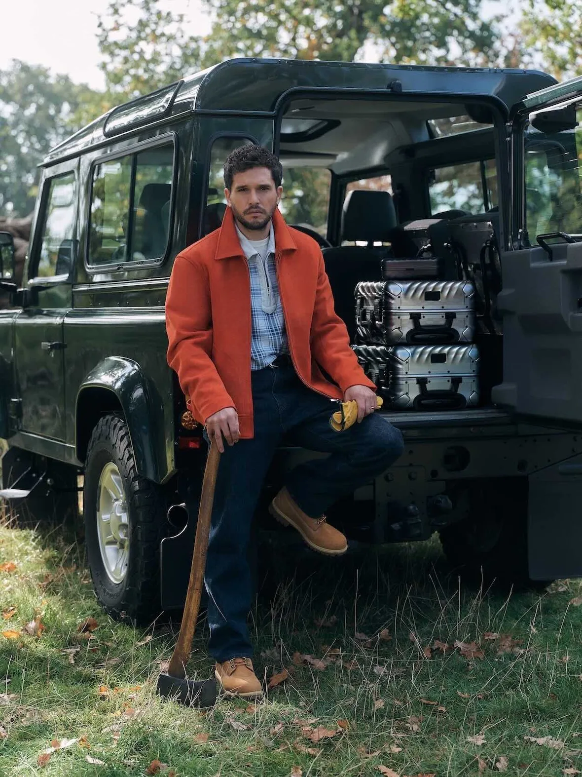 On-model, orange jacket, plaid shirt and tan lug-sole boots, leaning on axe by green 4x4 with silver suitcases, Winter 2026