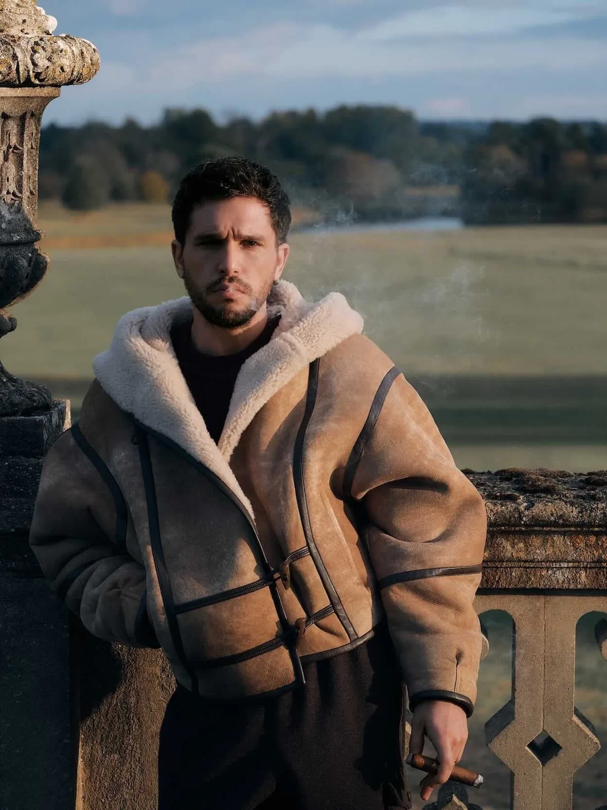 On-model close-up, tan shearling hooded jacket with dark leather trim and toggles, holding cigar, stone urn, Winter 2026