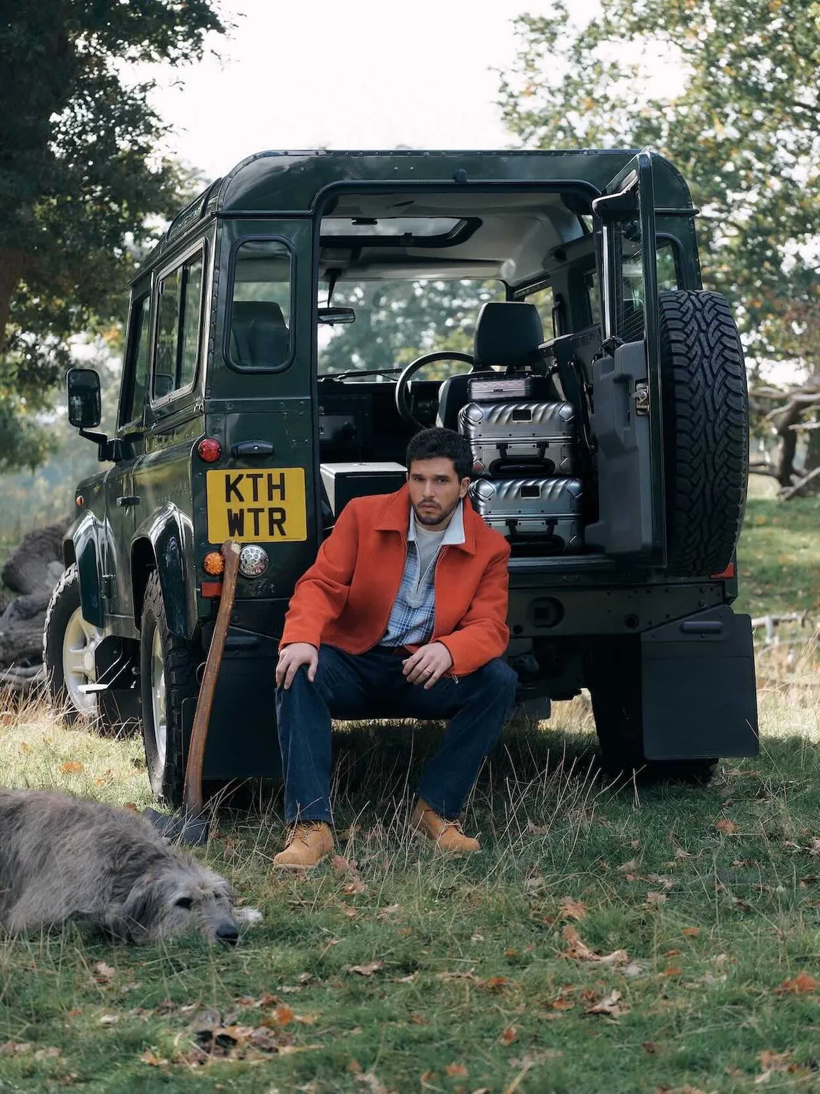 Man seated on rear of dark green 4x4, orange jacket, tan boots, yellow plate reading KTH WTR, dog on grass, Winter 2026